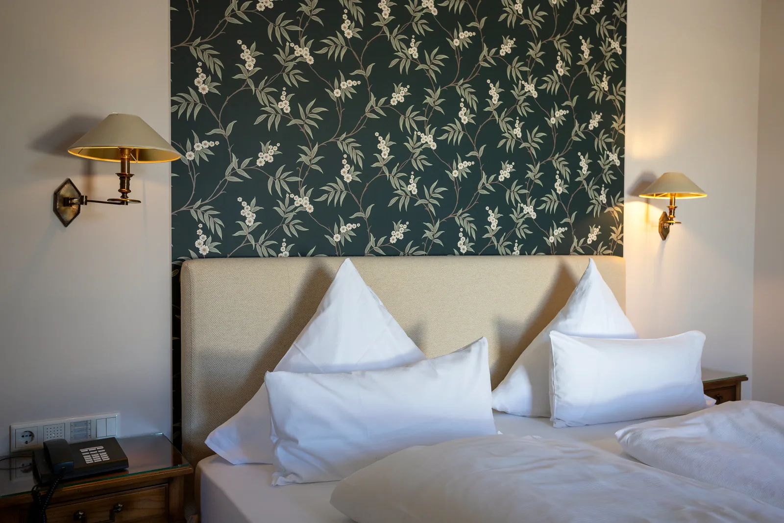 Weiße Kissen vor grüner Blumentapete mit Messing-Wandleuchte im Hotel Krone