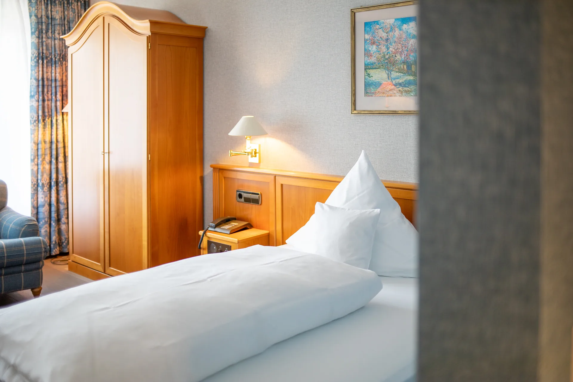 Einzelzimmer mit Holzschrank, Bett und Sessel mit blauem Muster