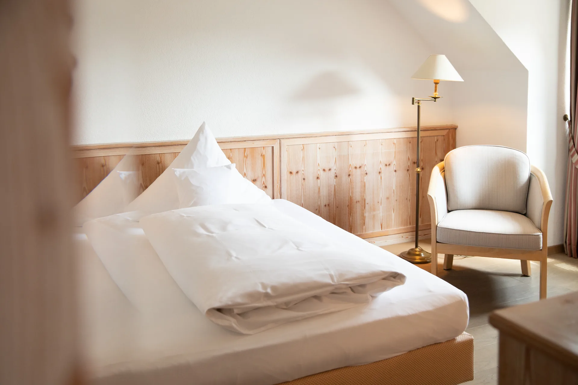 Einzelzimmer mit Bett, Holzvertäfelung und hellem Sessel am Fenster