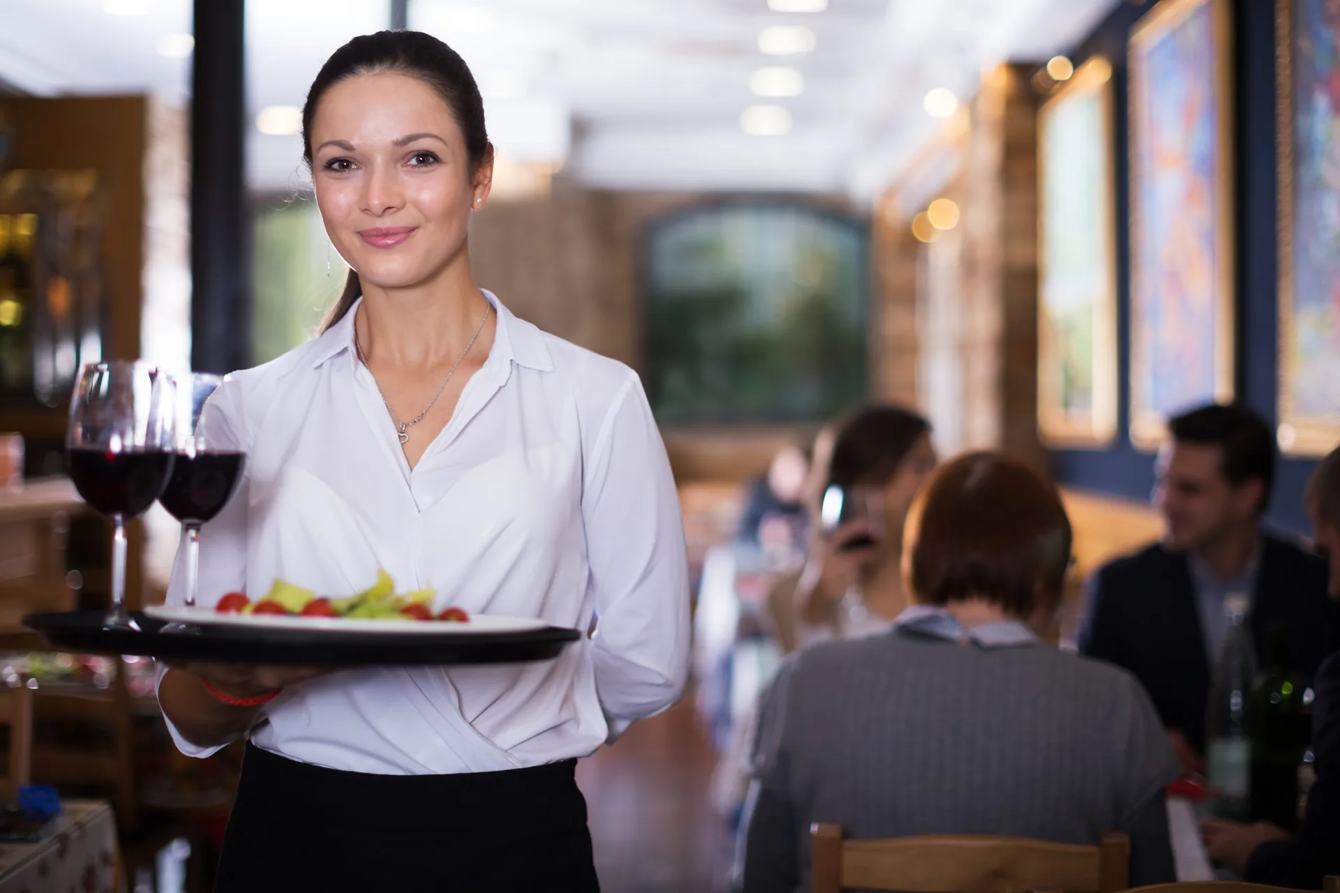 Servicemitarbeiterin trägt ein Tablett mit Rotweingläsern und einem Teller durch das Restaurant