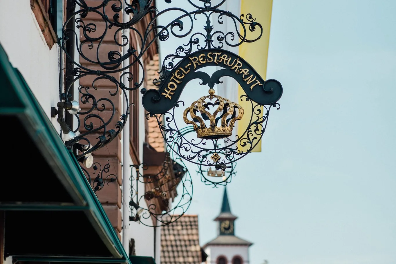 Schmiedeeisernes Wirtshausschild des Hotel Krone, im Hintergrund der Kirchturm von Herxheim-Hayna